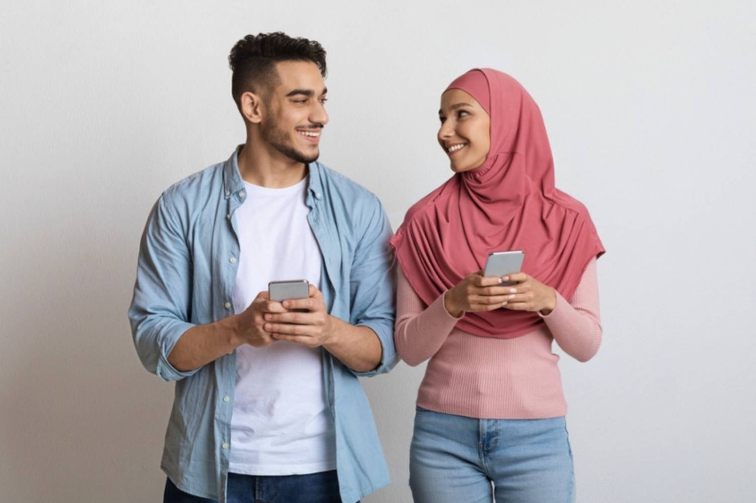 A man in a blue button up and woman in a pink shirt and hijab looking at each other admiringly with their phones in their hands