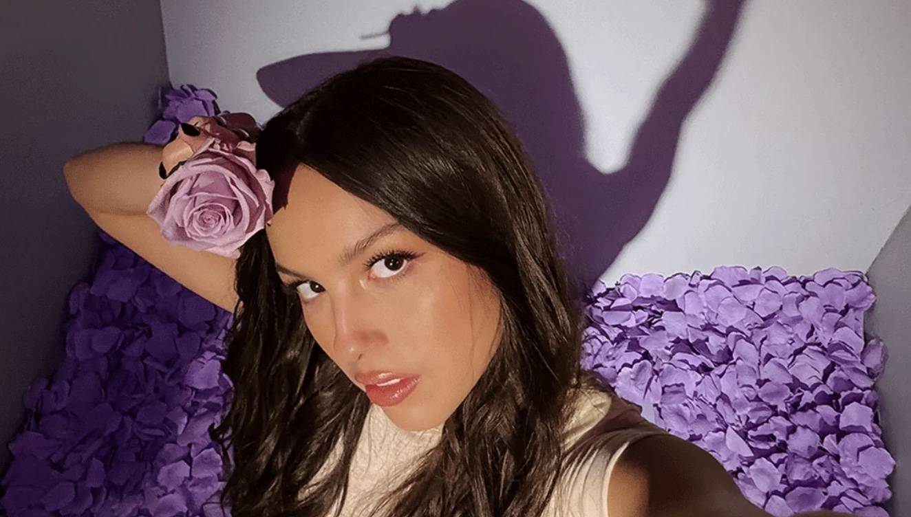 Olivia Rodrigo holding a purple rose sitting on purple rose petals