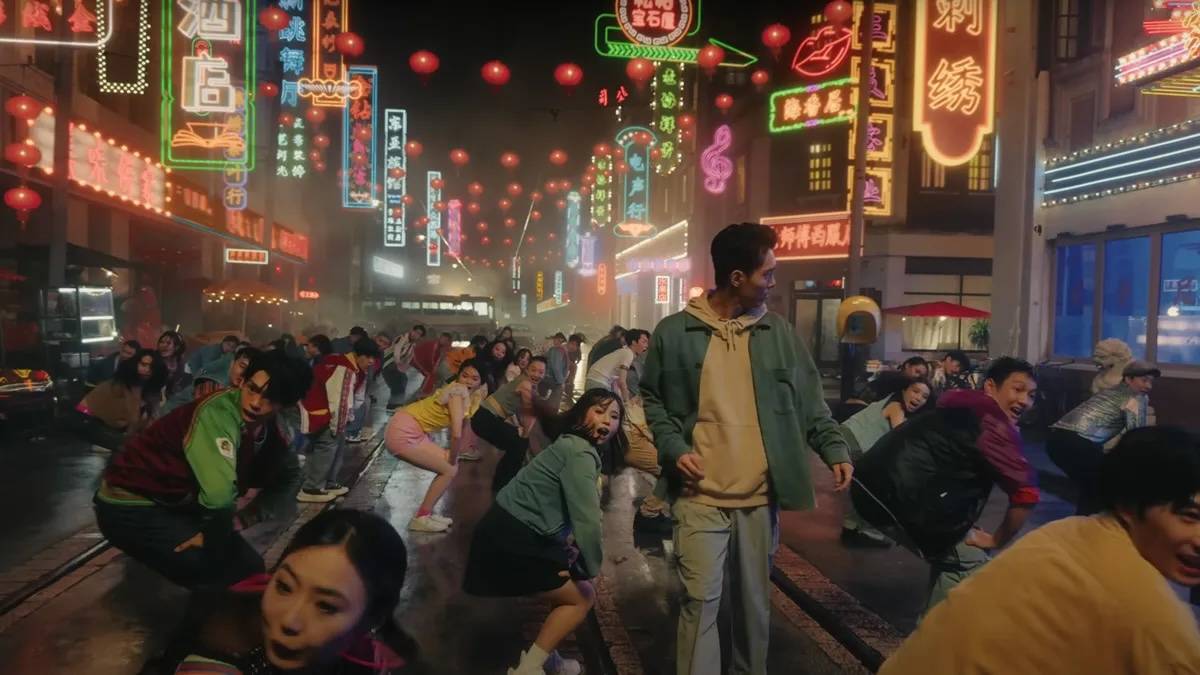 A man walks through a street lit with neon signs and filled with a dancing crowd.